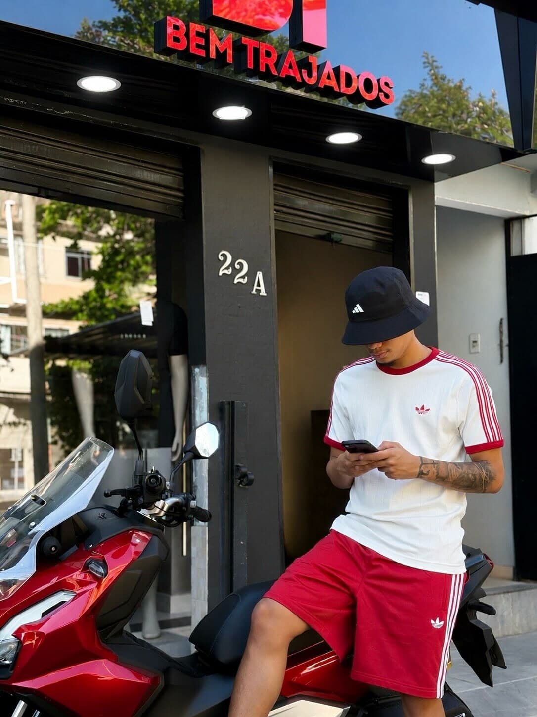 Cria de bucket Adidas e conjunto vermelho, sentado na moto vermelha, na frente da loja Bem Trajados, no Jacaré