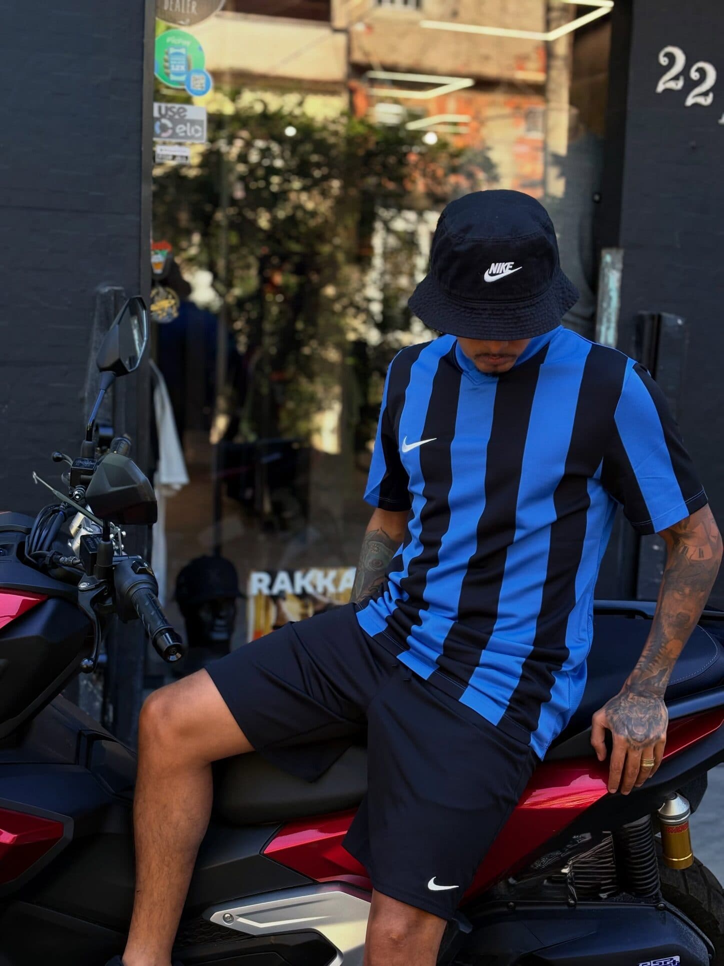 Cria de camisa listrada azul e preta e bucket Nike, sentado na moto na frente da loja, Rua do Rio 22A